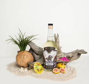 Image of a bottle of Ranch Rider Spirits Coconut Guava-infused Blanco Tequila with a blue, white, and gold label. At the top of the label, the Ranch Rider Spirits Co. logo and an illustration of a cowboy riding a horse. The label says “Coconut Guava Tequila, Blanco tequila infused with real fruit and all natural flavors. 60 Proof, 750 ml, 30% ABV.” The bottle is surrounded with sand, driftwood, lemons, and tiny paper umbrellas.