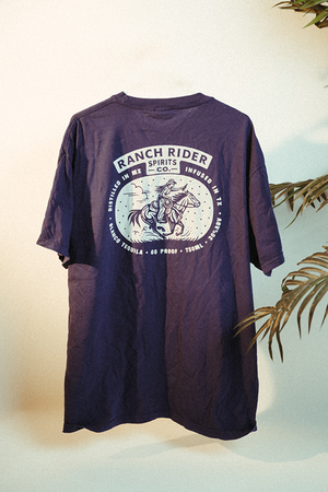 Image of a purple t-shirt with the Ranch Rider logo on the back, and below that an image of a cowboy on a horse with a rope on a desert backdrop in cream.