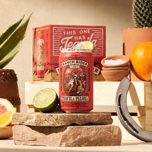 An image of the Ranch Rider Tequila Paloma pre-made cocktail can. The can is dark peach with dark red, cream, and yellow details. The label says "A premium tequila seltzer" at the top. Below that, there is an image of a cowboy on a horse with a rope on a desert backdrop. Below the logo, the label says: "Tequila Paloma, with only reposado tequila, sparkling water, sea salt, real grapefruit, lime and orange." The can is surrounded with rocks, succulents, grapefruit slices, and other western ephemera.