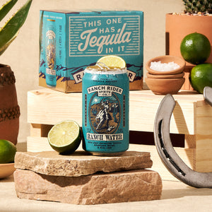 An image of the Original Ranch Water pre-made cocktail can. The can is turquoise with blue and gold details. The label says "A premium tequila seltzer" at the top. Below that, there is an image of a cowboy on a horse with a rope on a desert backdrop. Below the logo, the label says: "Ranch Water, with only reposado tequila, sparkling water, and real lime." The can has salt and lime on the rim and is surrounded with rocks, driftwood, limes, salt, and other western ephemera.