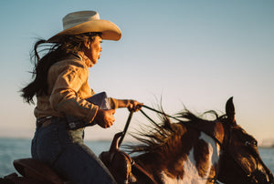 Image of a woman in western wear and a cowboy hat. She’s riding a brown and white horse on a beach.