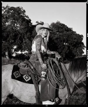 A black and white image of a blond woman in western wear and a white hat. She’s holding a bottle of tequila and sitting on top of a horse with a saddle. Across the image is the Ranch Rider Spirits Co. logo in white.