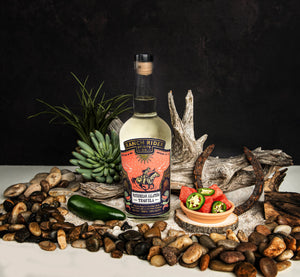 Image of a bottle of Ranch Rider Spirits Watermelon Jalapeño-infused Blanco Tequila with a blue, pink, and gold label. At the top of the label, the Ranch Rider Spirits Co. logo and an illustration of a cowboy riding a horse. The label says “Watermelon Jalapeño Tequila, Blanco tequila infused with real fruit and all natural flavors. 60 Proof, 750 ml, 30% ABV.” The bottle is surrounded with rocks, driftwood, jalapeños, and watermelon.