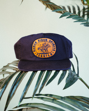 Dark blue baseball cap with a yellow patch that says "Ranch Rider Spirits Tequila" around an illustration of a rider on a horse.