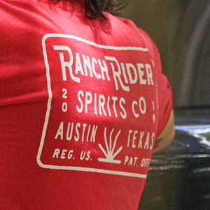 Image of a red t-shirt with the words “Ranch Rider Spirits Co., Austin, Texas” and an agave icon in cream on the back. The shirt is available for purchase on the Dry Goods page.