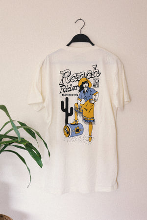 Image of a white t-shirt with a dark blue, yellow, and light blue design. The back of the t-shirt has a cowgirl standing in western wear with her foot up on a can and a cactus in the background. Behind the woman it says, “Ranch Rider Spirits, Tex Mex.”