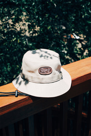 White bucket hat with a brown and white embroidered design that says “Buck it” in script lettering.